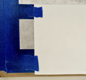 Photograph of a blank white rectangular sheet taped onto a surface with three pieces of blue painter's tape. The background consists of vertical gray and blue stripes with a brown edge visible on the right side.
