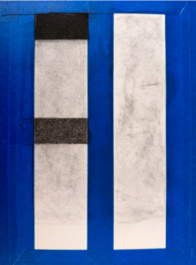 Photograph of a white surface with a rectangular area in the center shaded dark gray and bordered by blue painter's tape. The tape forms an incomplete frame around the shaded area, with some pieces overlapping and edges uneven.