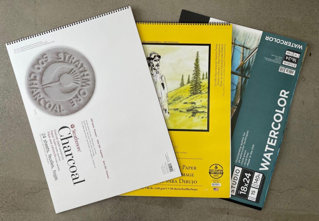 Photograph of three art paper pads arranged on a gray surface, including a white charcoal paper pad, a yellow drawing paper pad with an illustration of a person and trees, and a blue watercolor paper pad labeled "WATERCOLOR." Each pad displays branding, size, and paper type information, highlighting options for different artistic mediums.