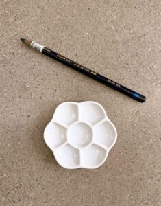Photograph showing a black Derwent Inktense pencil and a white mixing palette with six petal-shaped wells arranged around a central circular well. Items are placed on a textured beige surface, highlighting tools commonly used for drawing and painting.