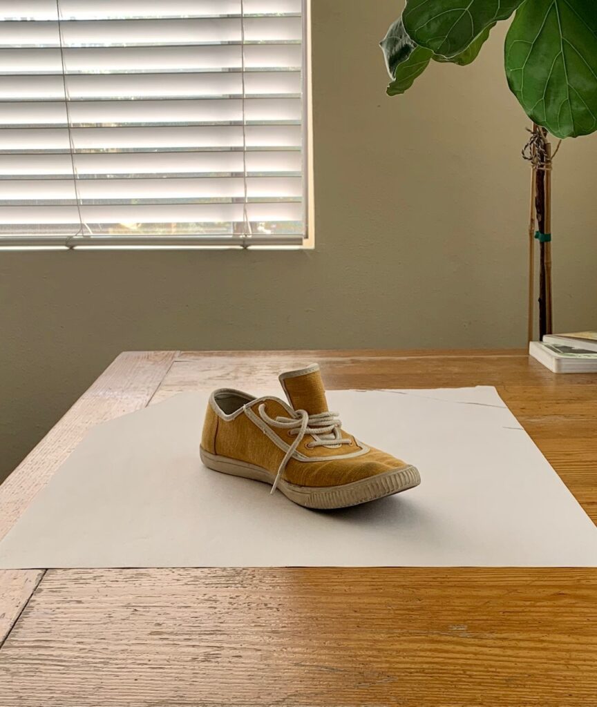 Photograph of a single yellow canvas sneaker with white laces placed on a white sheet of paper on a wooden table.