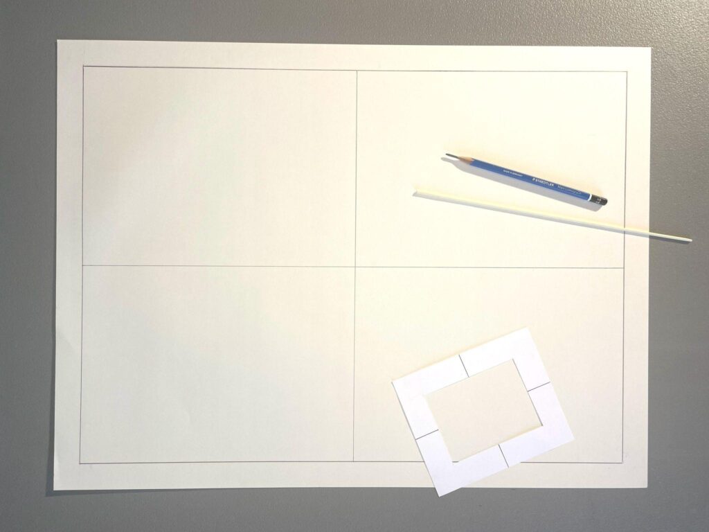 Photograph of a blank white sheet divided into four rectangular sections by thin lines, placed on a gray surface. A blue pencil, a white stick, and a small white rectangular viewfinder frame with cut-out sections rest on the sheet, suggesting preparation for a drawing project.