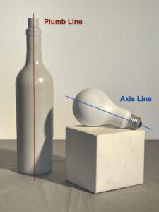 Photograph showing a grey bottle standing upright with a red vertical "Plumb Line" and a white light bulb lying on its side on a white cube with a blue diagonal "Axis Line." The image illustrates concepts of vertical alignment and axis orientation using labeled colored lines for clarity.