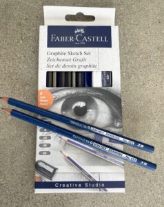 Photograph of a Faber-Castell Graphite Sketch Set packaging containing eight drawing pencils and a sharpener. Packaging features a detailed eye sketch, pencil hardness labels (6B, 4B, 2B, B), and two blue pencils labeled 2B and 3H placed on top.