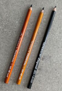 Photograph of three drawing pencils arranged diagonally on a gray textured surface. Pencils include two brown General's charcoal pencils labeled 557-29 med and 557-48 soft, and one black General's lacquer pencil labeled 555.