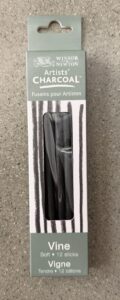 Photograph of a rectangular package of Winsor & Newton Artists' Charcoal labeled "Vine Soft 12 sticks." Package features a gray and white striped design with a clear window showing black charcoal sticks inside.