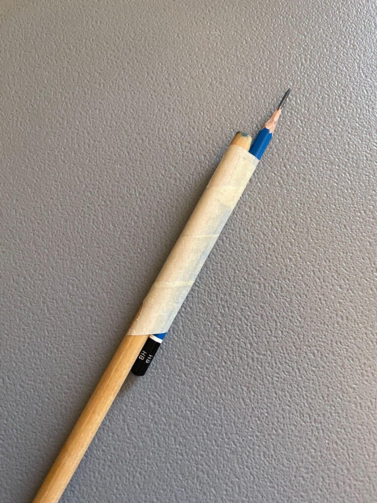 Photograph of a makeshift tool created by taping a sharpened pencil to a wooden stick, likely for extended reach or precision. The pencil is blue with a visible "HB" label, and the tape is beige, securing the pencil firmly to the stick against a textured gray background.