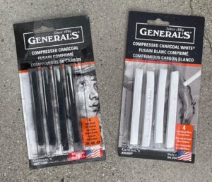 Photograph of two packages of General's compressed charcoal sticks, one containing black charcoal in varying hardness levels (2B, 4B, 6B) and the other containing white charcoal sticks. Both packages feature black and white designs with product details in English, French, and Spanish, and display an American flag indicating they are made in the USA.