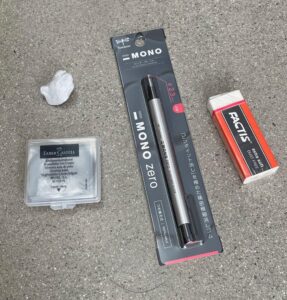 Photograph showing four different erasers arranged on a gray speckled surface, including a white kneaded eraser, a Faber-Castell kneaded eraser in a clear case, a Tombow MONO Zero precision eraser in black and silver packaging, and a red and white FACTIS eraser.