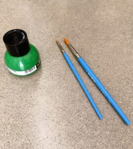 Photograph showing two blue-handled paintbrushes and a small green paint bottle with a black cap on a speckled gray surface. The paintbrushes vary in size.