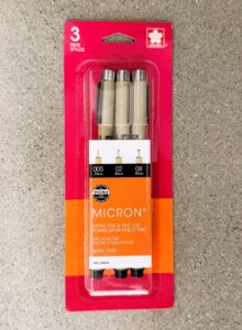 Photograph of a three-pack of Sakura Pigma Micron pens in original red and orange packaging, highlighting pen tip sizes 005 (0.20mm), 02 (0.30mm), and 08 (0.50mm). Packaging emphasizes extra fine and fine line archival ink in black.