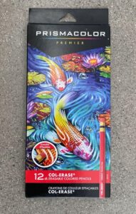 Photograph of a Prismacolor Premier colored pencil box featuring vibrant artwork of two koi fish swimming among lily pads in a blue pond. The packaging highlights 12 erasable colored pencils with a red label at the bottom indicating "Col-Erase" and includes an image of a pencil eraser.