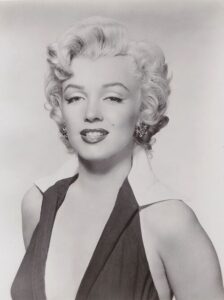Black and white photograph of a woman with styled blonde hair, wearing a dark sleeveless dress with a plunging neckline and light-colored collar. The portrait captures a confident expression with subtle makeup, including dark lipstick and earrings.