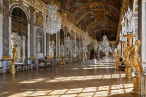 Photograph of an opulent palace hall featuring a long corridor with large arched mirrors, ornate gold statues, and multiple crystal chandeliers. The ceiling is richly decorated with intricate paintings, and sunlight casts patterned shadows on the polished wooden floor.