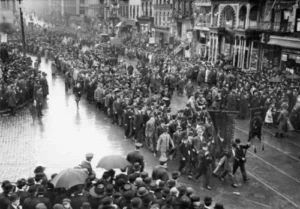 A large parade with marchers and spectators on a cobblestone street on a rainy day.