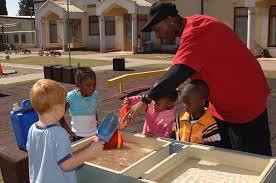 A caregiver assists five children of various backgrounds in a water play activity outdoors, filled with joy and engagement in a sunny playground