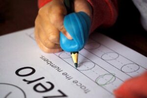 A child's hand holding a blue pencil gripper traces a circle on a worksheet with outlined shapes.