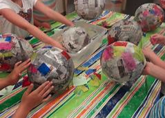 Children crafting papier-mâché balloons with newspaper and colorful tissue paper on a striped tablecloth.