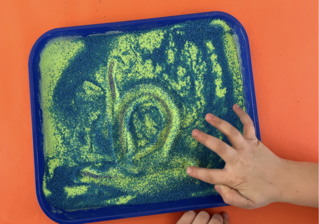 Child drawing a spiral in green and blue sand on a blue tray.