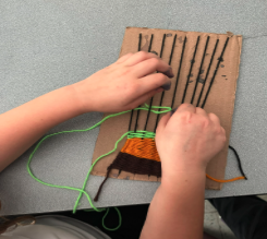 Hands weaving orange and green yarn on a cardboard loom.
