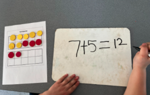 Double 10 frame with 7 yellow dots, 5 red dots and a child writing 7+5=12 on white board