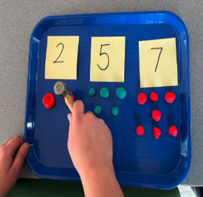 Tray with the numbers 2, 5 and 7 on sticky notes with balls of clay and child's hand using a hammer to press down clay balls