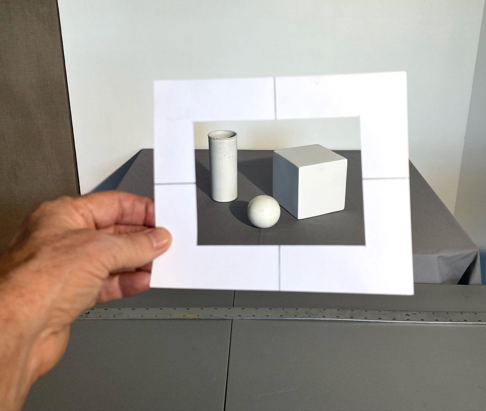Photograph showing a hand holding a white rectangular viewfinder frame with cutouts, focusing on three white geometric shapes—a cylinder, sphere, and cube—arranged on a gray surface. The frame highlights the shapes' shadows and lighting, emphasizing their three-dimensional forms against a neutral background.