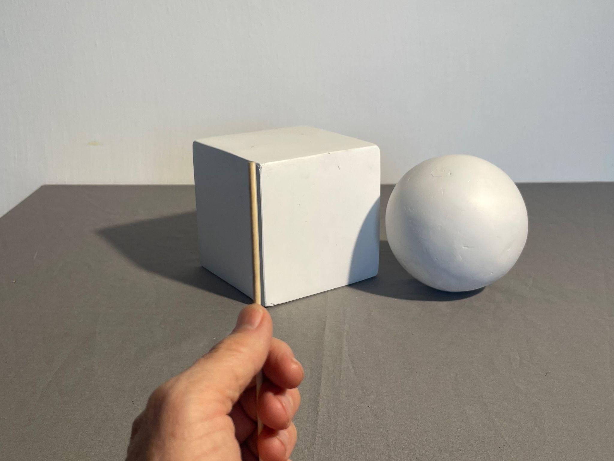 Photograph showing a white cube and a white sphere placed on a gray surface against a plain light background. A hand is positioned in the foreground, partially visible and holding a sighting tool vertically against the cube.