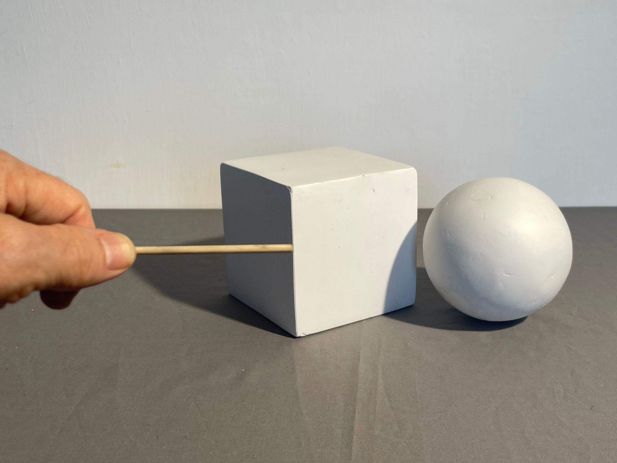Photograph showing a hand holding a wooden stick sighting tool horizontal to a white cube placed on a gray surface next to a white sphere.