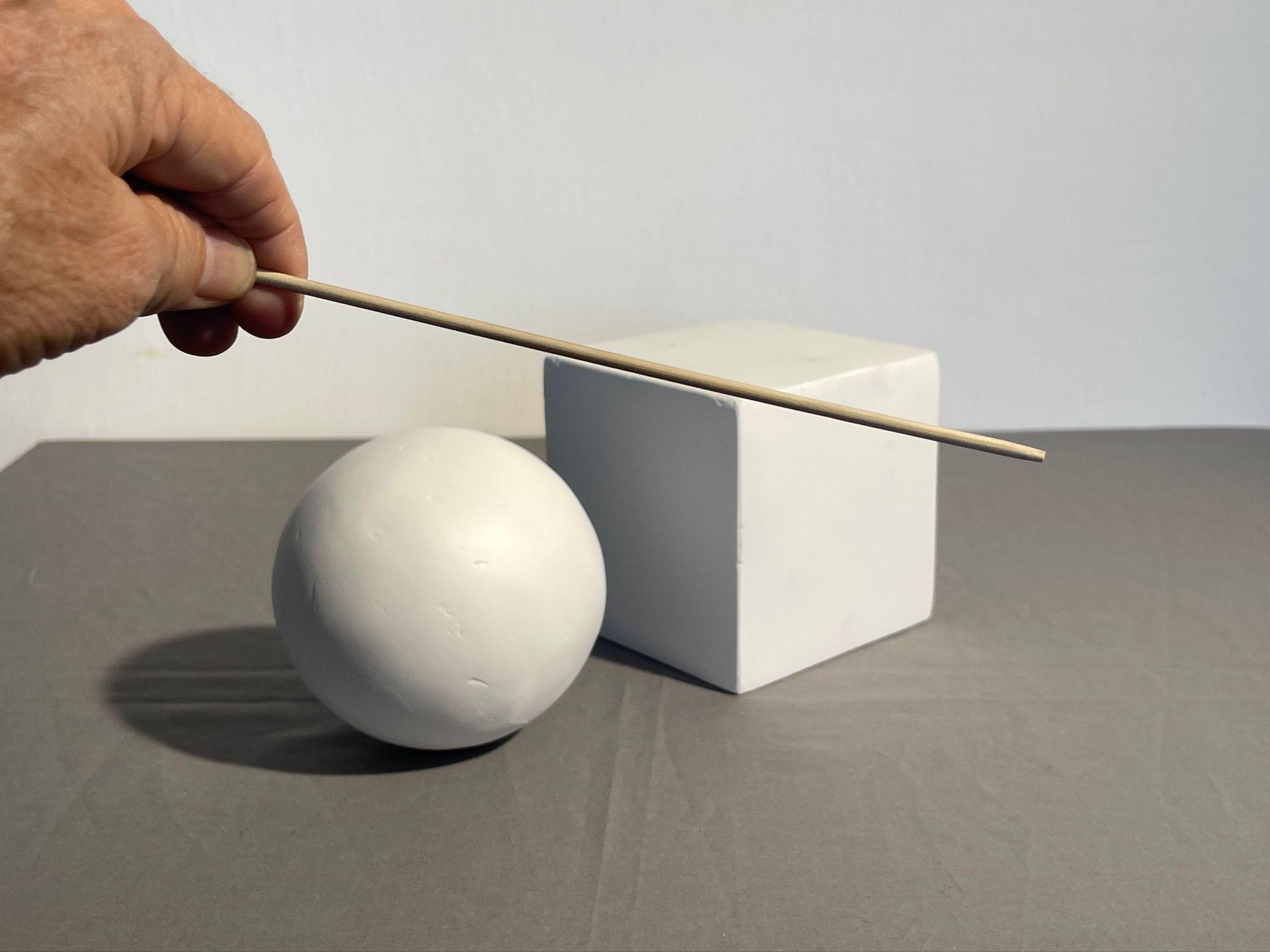 Photograph showing a hand holding a thin wooden stick as a sighting tool angled above a white sphere and a white cube placed on a gray surface. The setup highlights geometric shapes and spatial relationships between the sphere, cube, and stick.