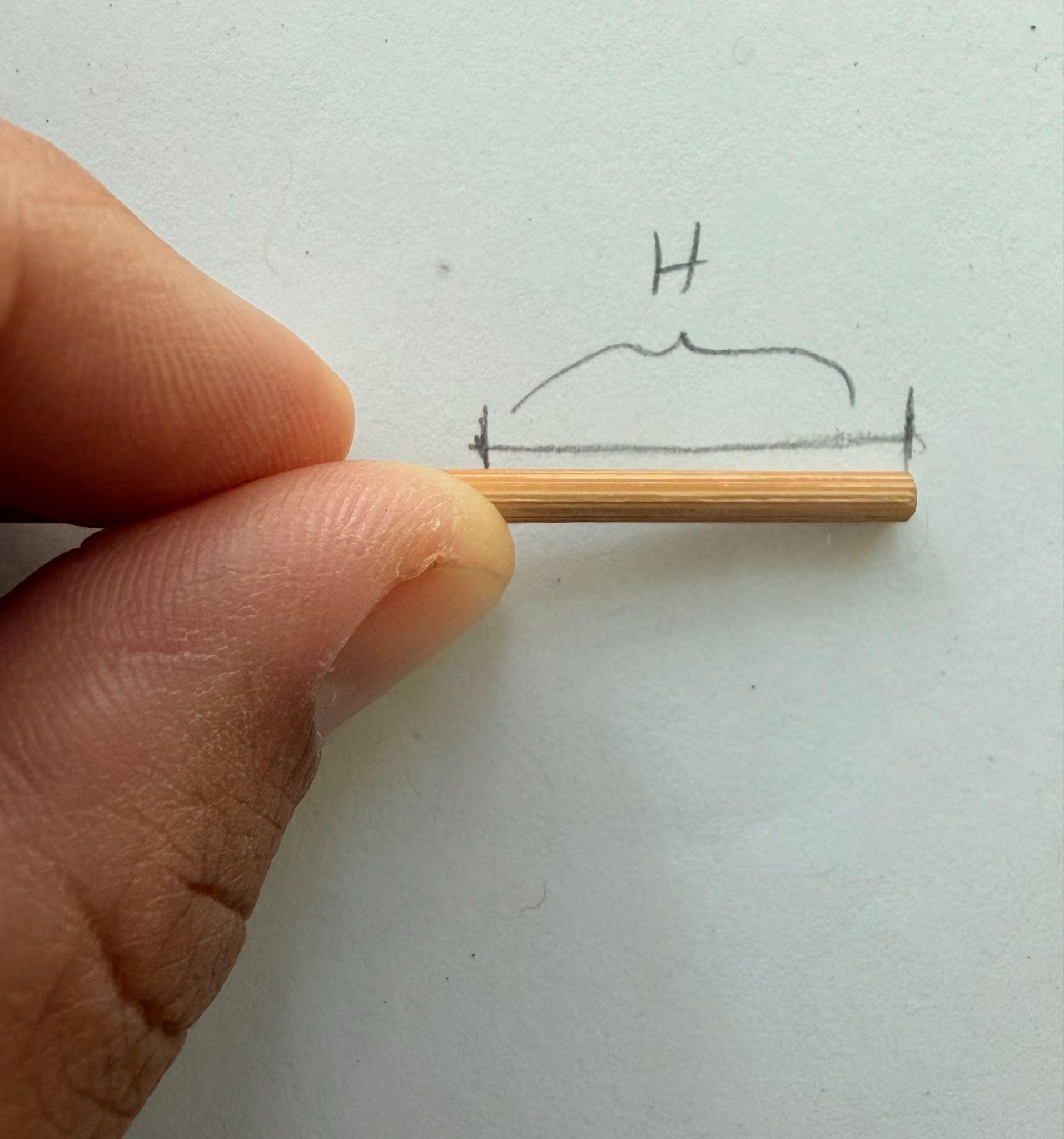 Photograph showing a close-up of a hand holding a small wooden stick horizontally against a white background with a hand-drawn bracket labeled "H" above the stick. The image highlights measurement using a sighting tool.