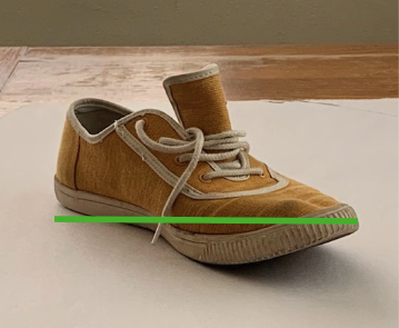 Photograph of a single brown canvas sneaker with white laces and a white rubber sole placed on a white surface against a wooden background. The shoe is positioned at an angle showing the side and front with a green horizontal line that denotes the width measurement annotation.