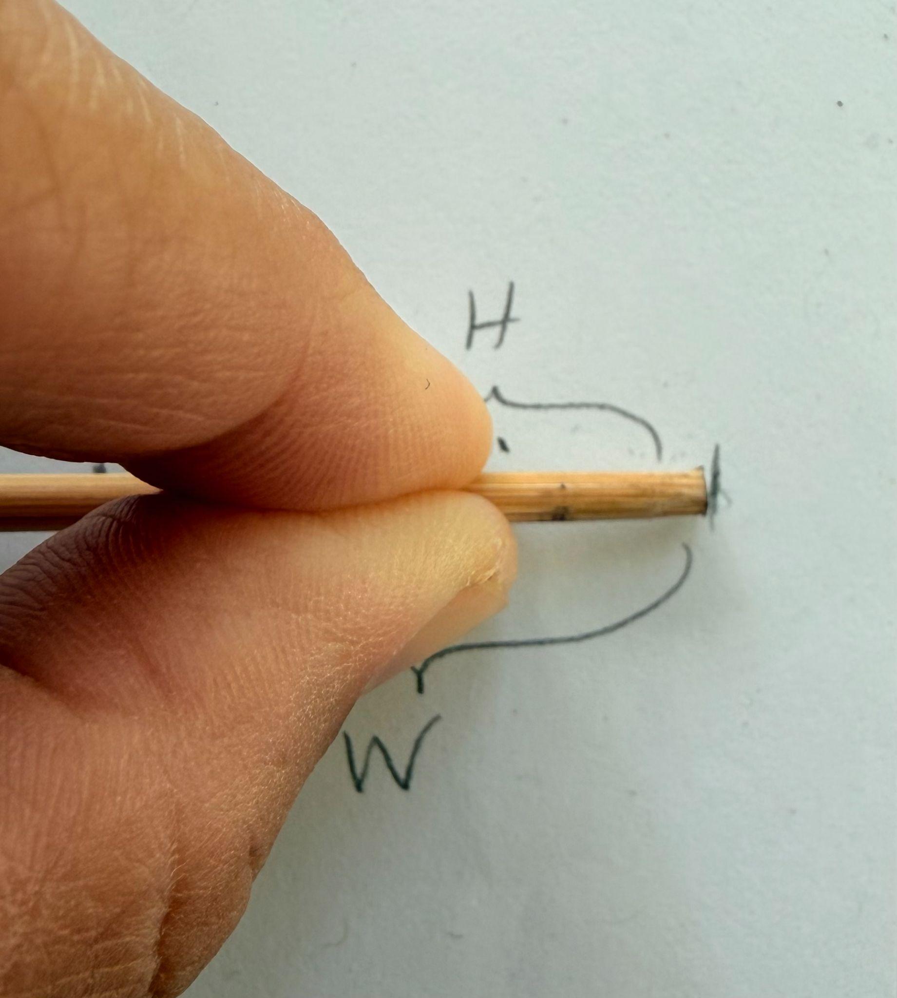 Photograph showing a close-up of a hand holding a wooden stick aligned with a small black mark on a white surface. Handwritten letters "H" and "W" are visible near the stick, indicating labeled measurements.