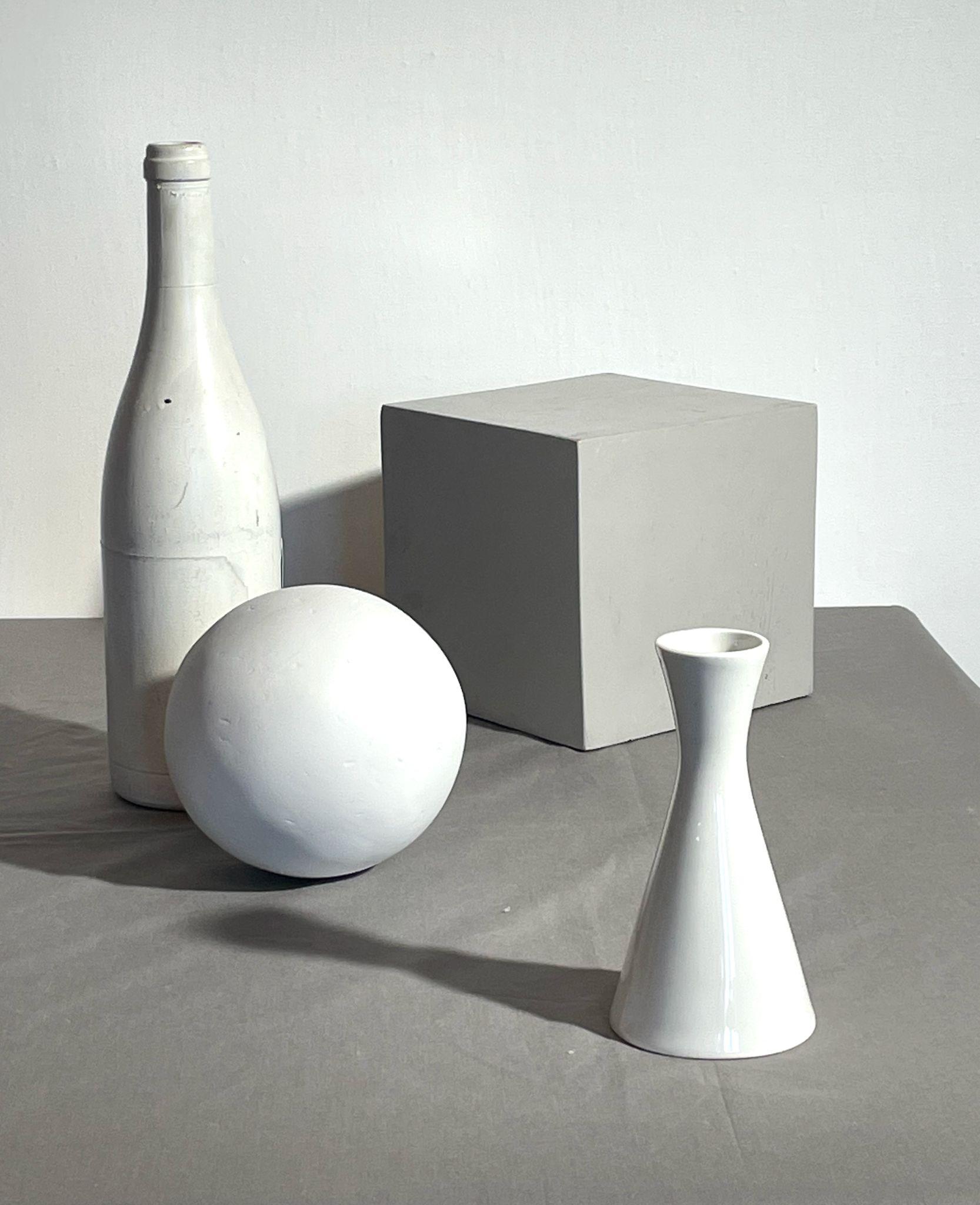 Photograph of four white geometric objects—a bottle, sphere, cube, and vase—arranged on a gray surface against a plain white background. The composition highlights overlapping shapes with shadows, emphasizing form and light interaction.