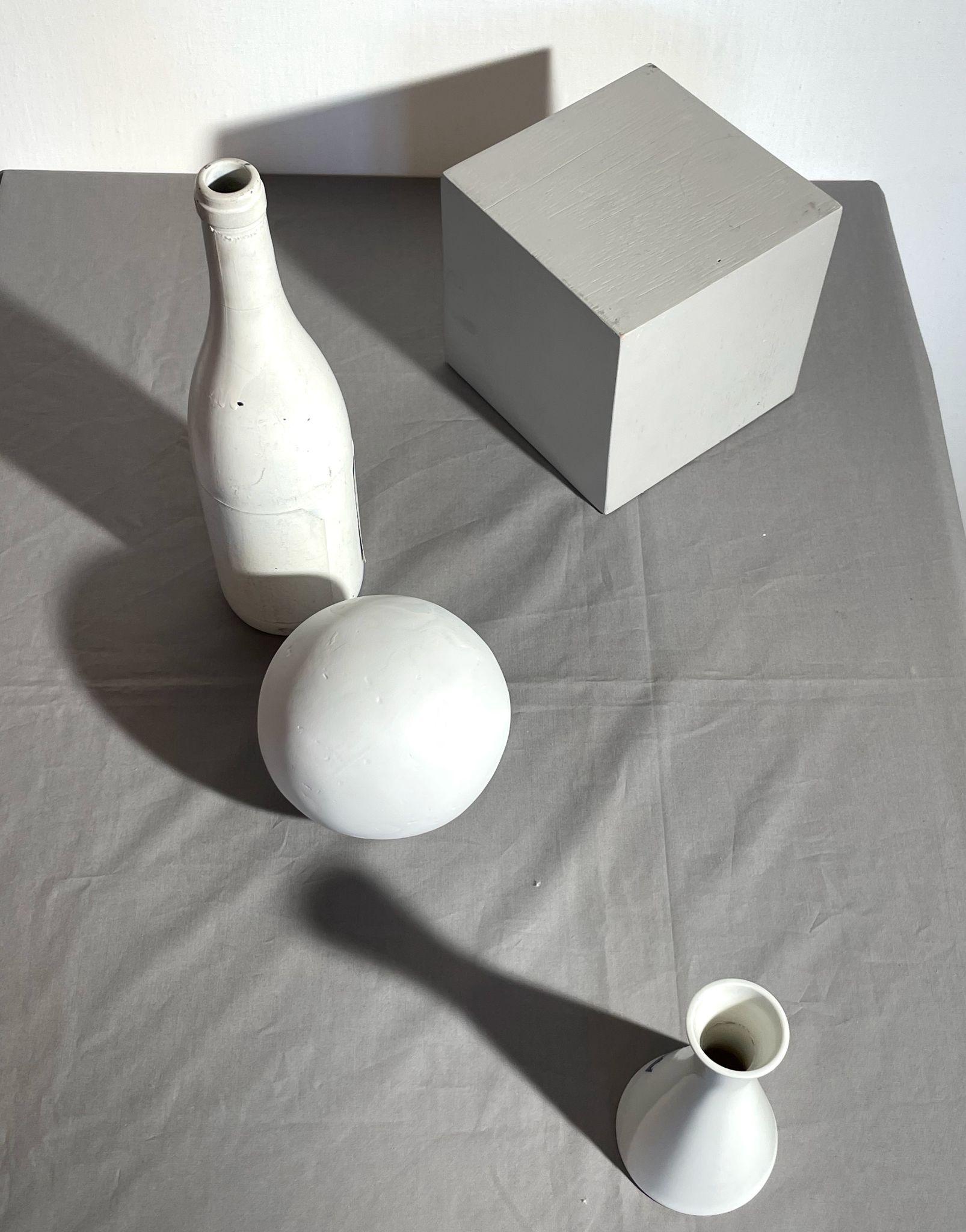 Photograph of four white geometric objects—a bottle, a sphere, a vase, and a cube—arranged on a gray fabric surface with distinct shadows cast by light from the upper left. The composition highlights the shapes from an angle above and looking down on the arrangemernt, emphasizing spatial relationships and lighting effects.