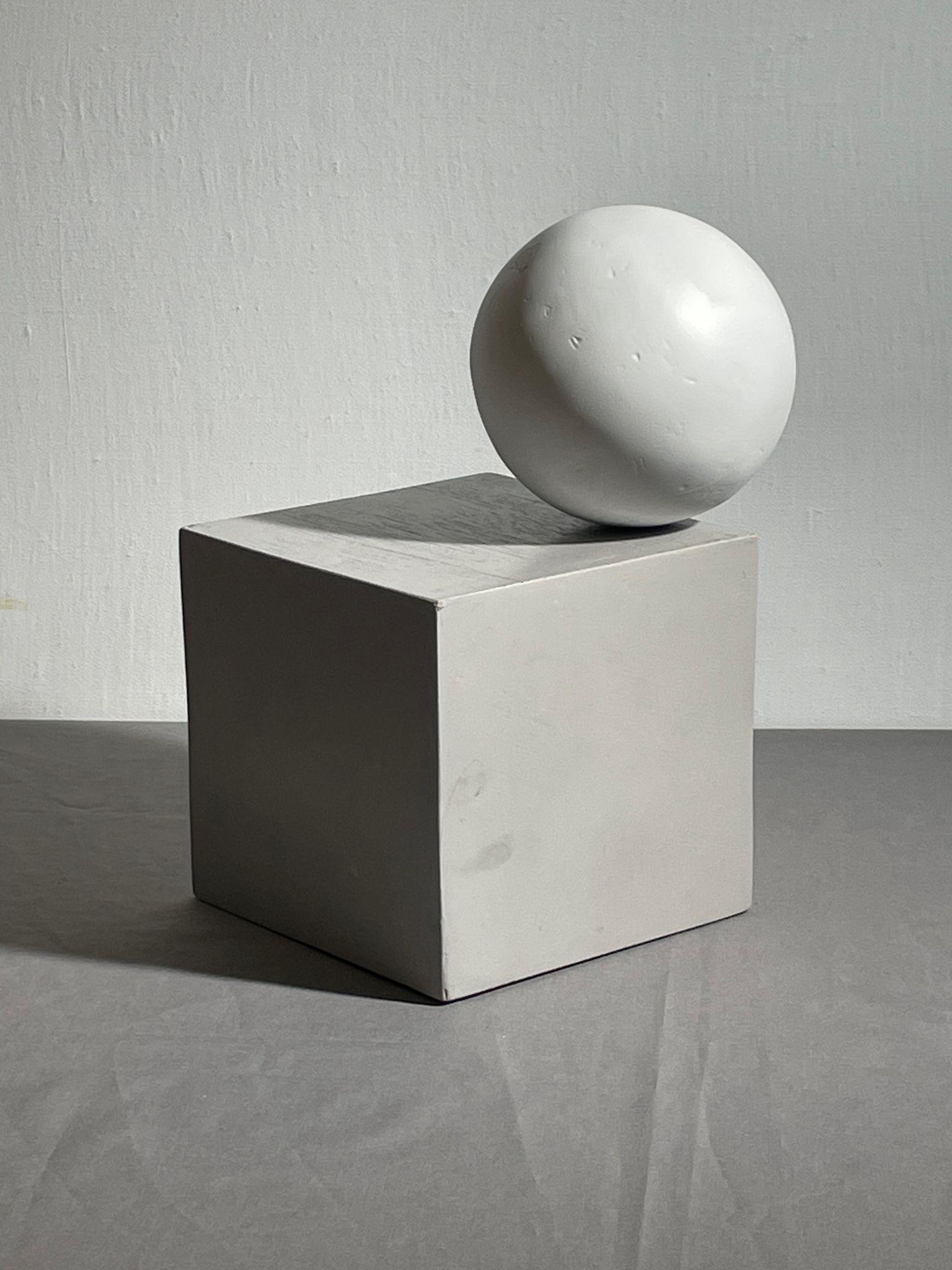 Photograph of a white sphere resting on a white cube against a plain wall background. Lighting creates distinct shadows on the cube and surface, emphasizing geometric shapes and texture.