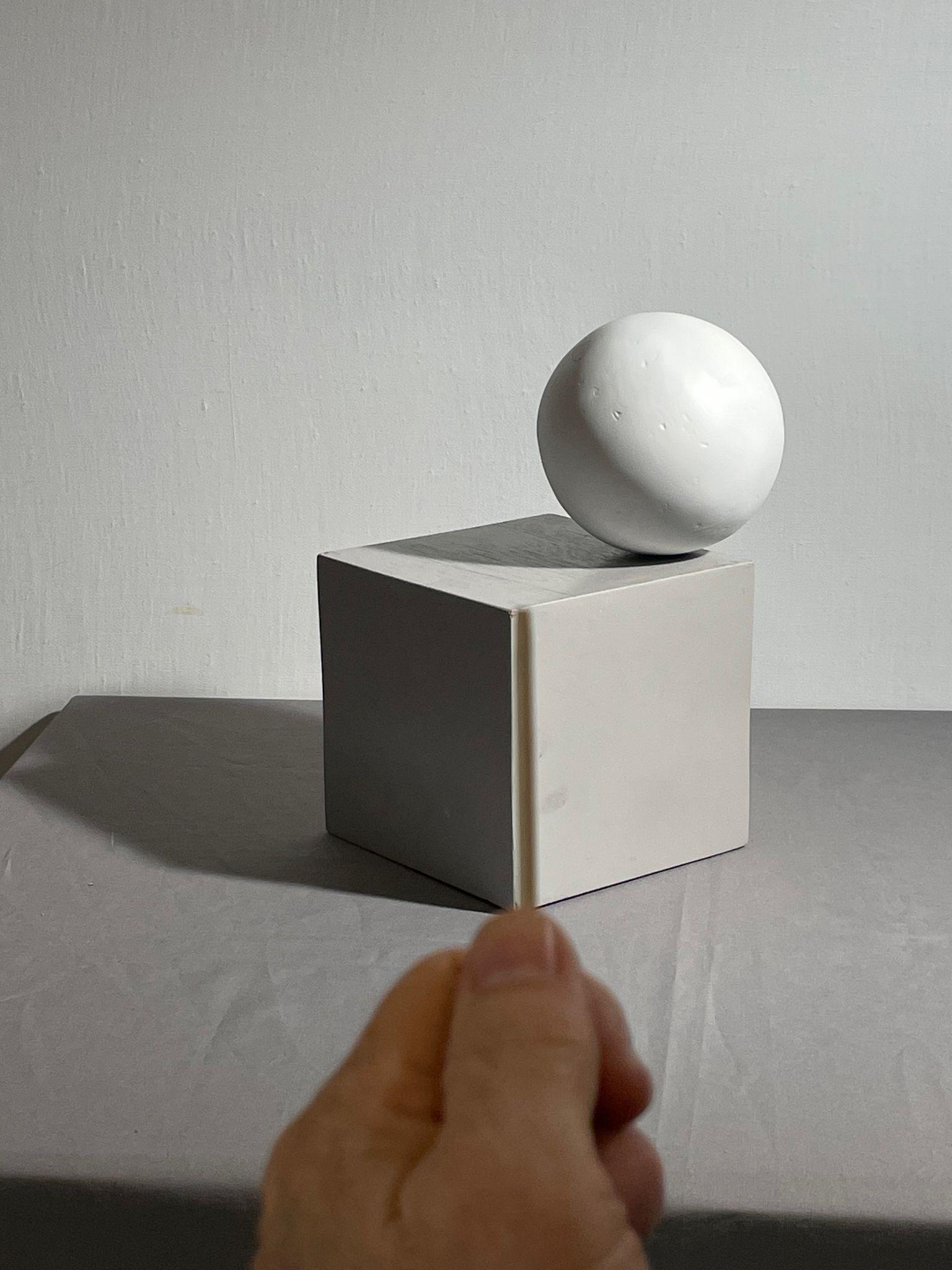Photograph of a white sphere resting on a white cube against a plain background, with a hand holding a small sighting tool vertically in the foreground. The composition highlights geometric shapes, shadows, and lighting contrasts.
