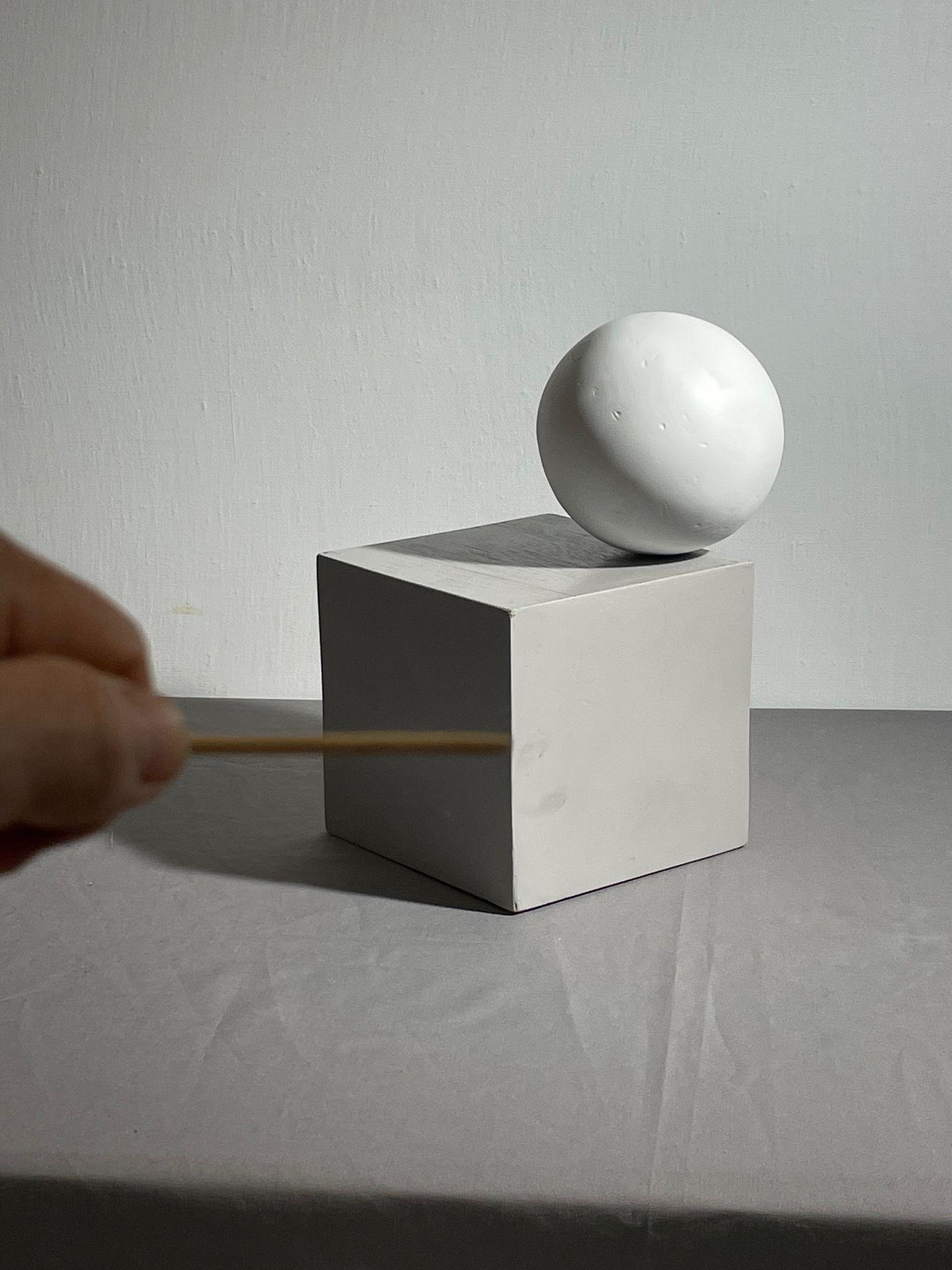 Photograph showing a white spherical object resting on a white cube against a plain gray background. A hand holding a thin sighting tool stick horizontally is positioned in the foreground.