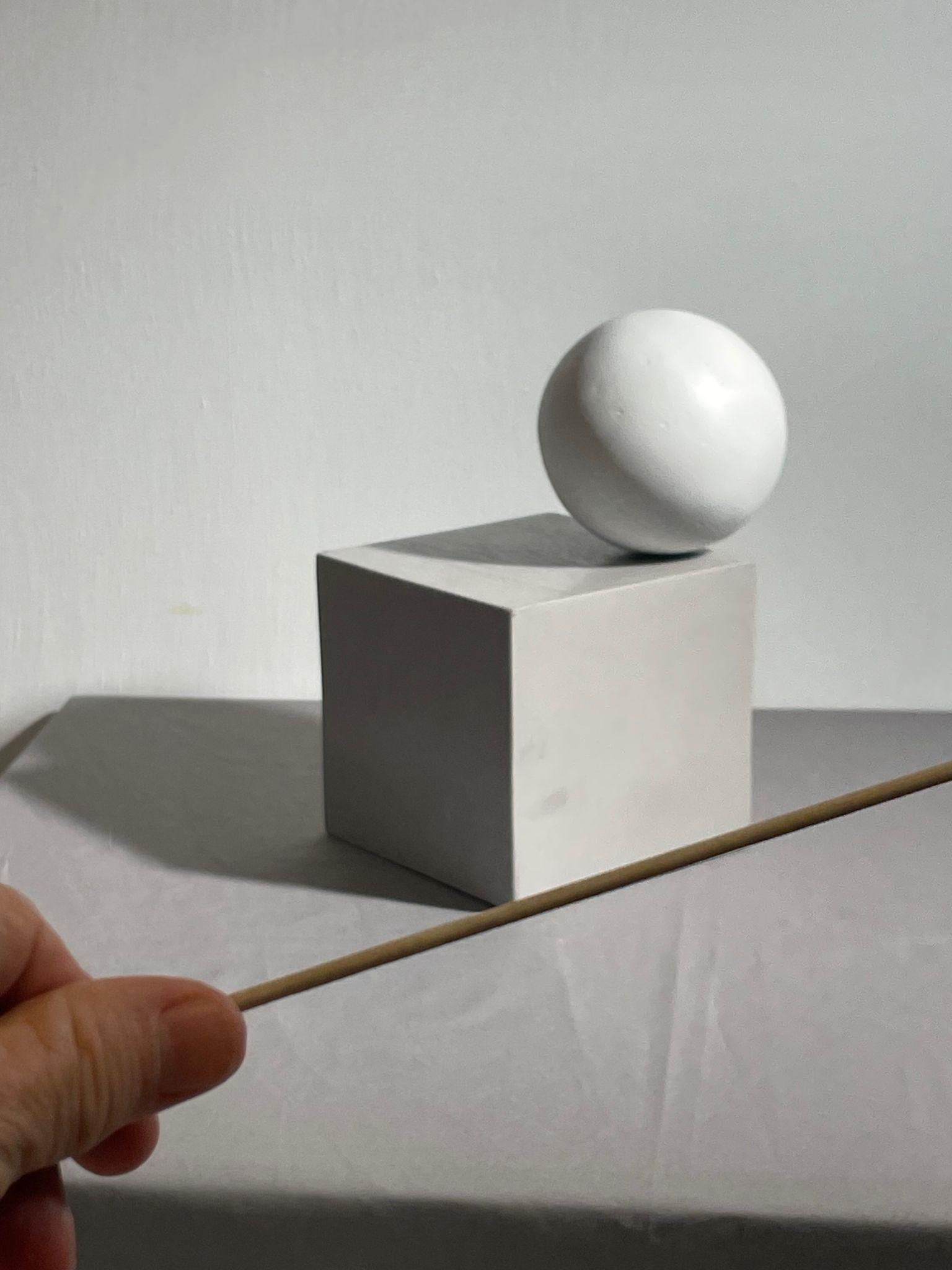 Photograph showing a white sphere balanced on a white cube against a plain background, with a hand holding a thin wooden stick angled diagonally against the bottom of the cube. The composition highlights the horizontal angle of the bottom edge of the cube.
