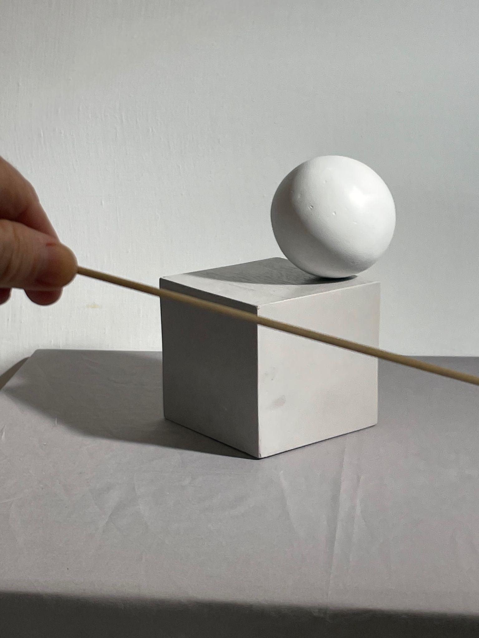 Photograph showing a white sphere balanced on a white cube placed on a gray surface with a plain light background. A hand holds a thin wooden stick diagonally across the upper part of the cube, emphasizing the horizontal angle of the top of the cube.