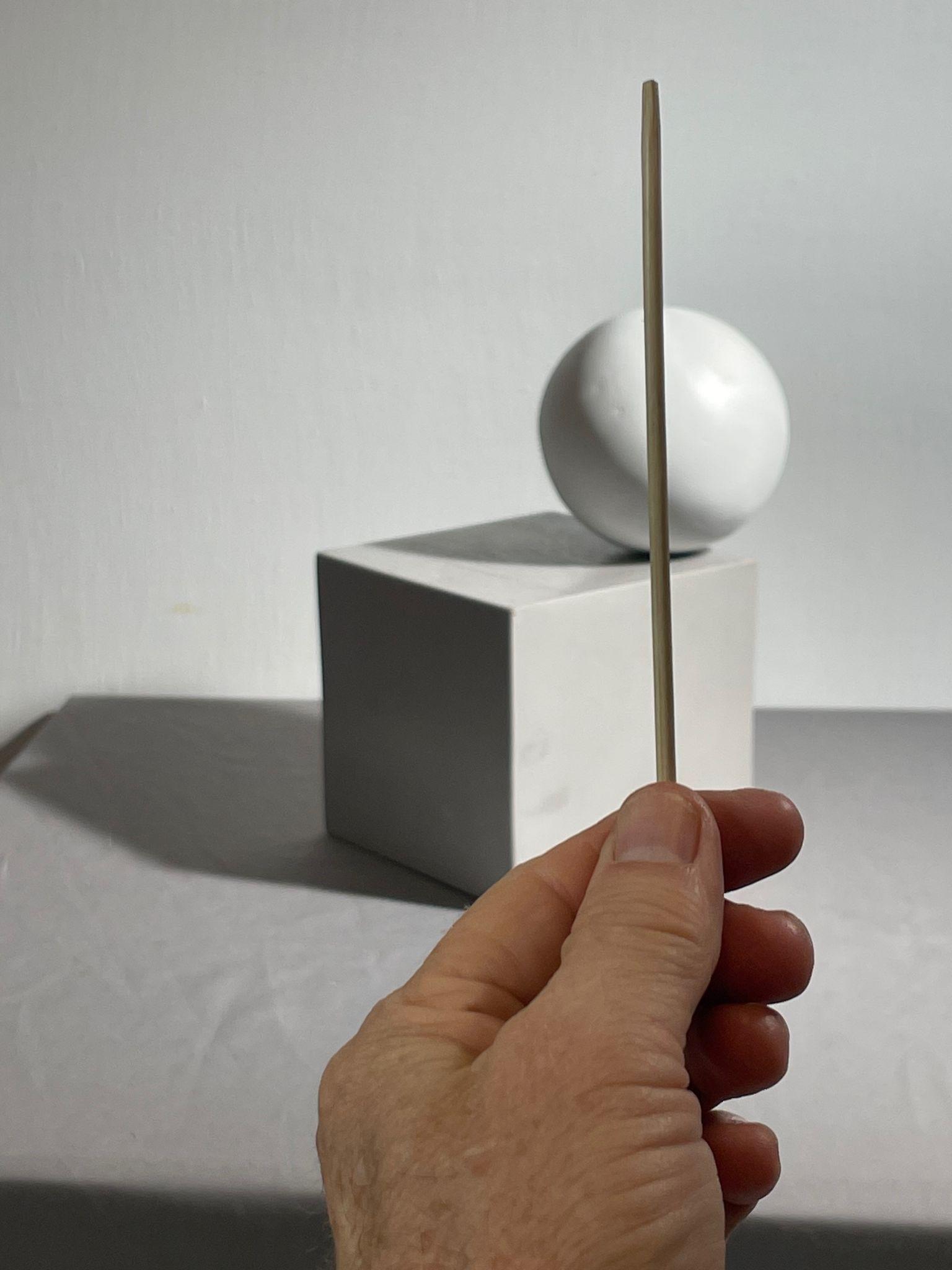 Photograph showing a hand holding a thin, vertical sighting stick in front of a white cube with a white sphere resting on top.