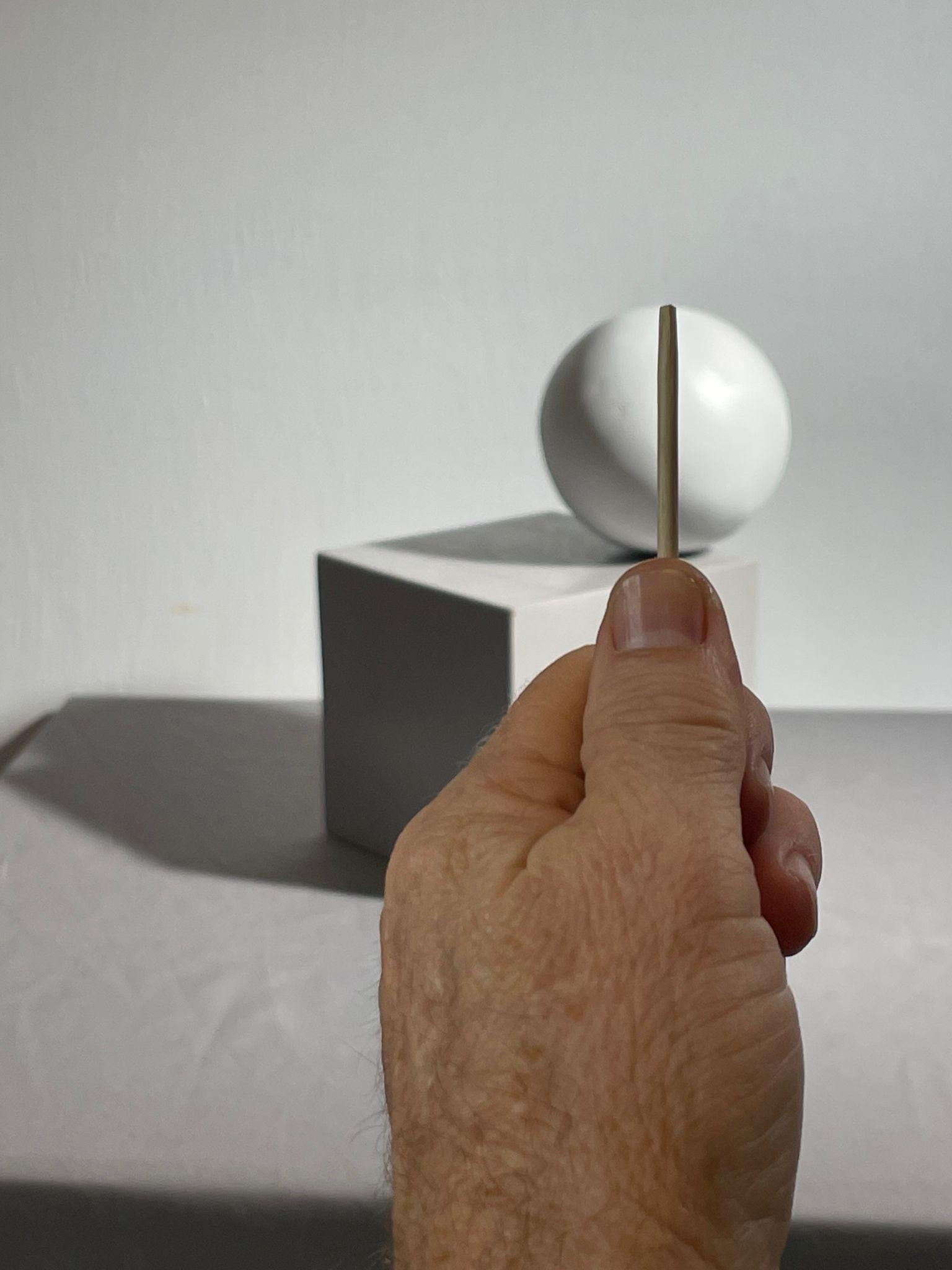Photograph showing a hand holding a thin vertical sighting tool, aligned with a white sphere resting on a gray cube in the background. The composition highlights perspective and alignment, with shadows cast on a plain surface and wall.