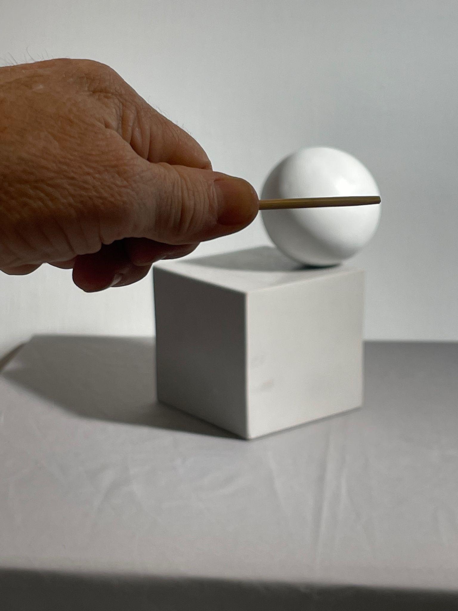 Photograph showing a hand holding a thin stick horizontally in front of a white sphere resting on a white cube, all set against a plain gray background. The composition highlights geometric shapes and shadows.