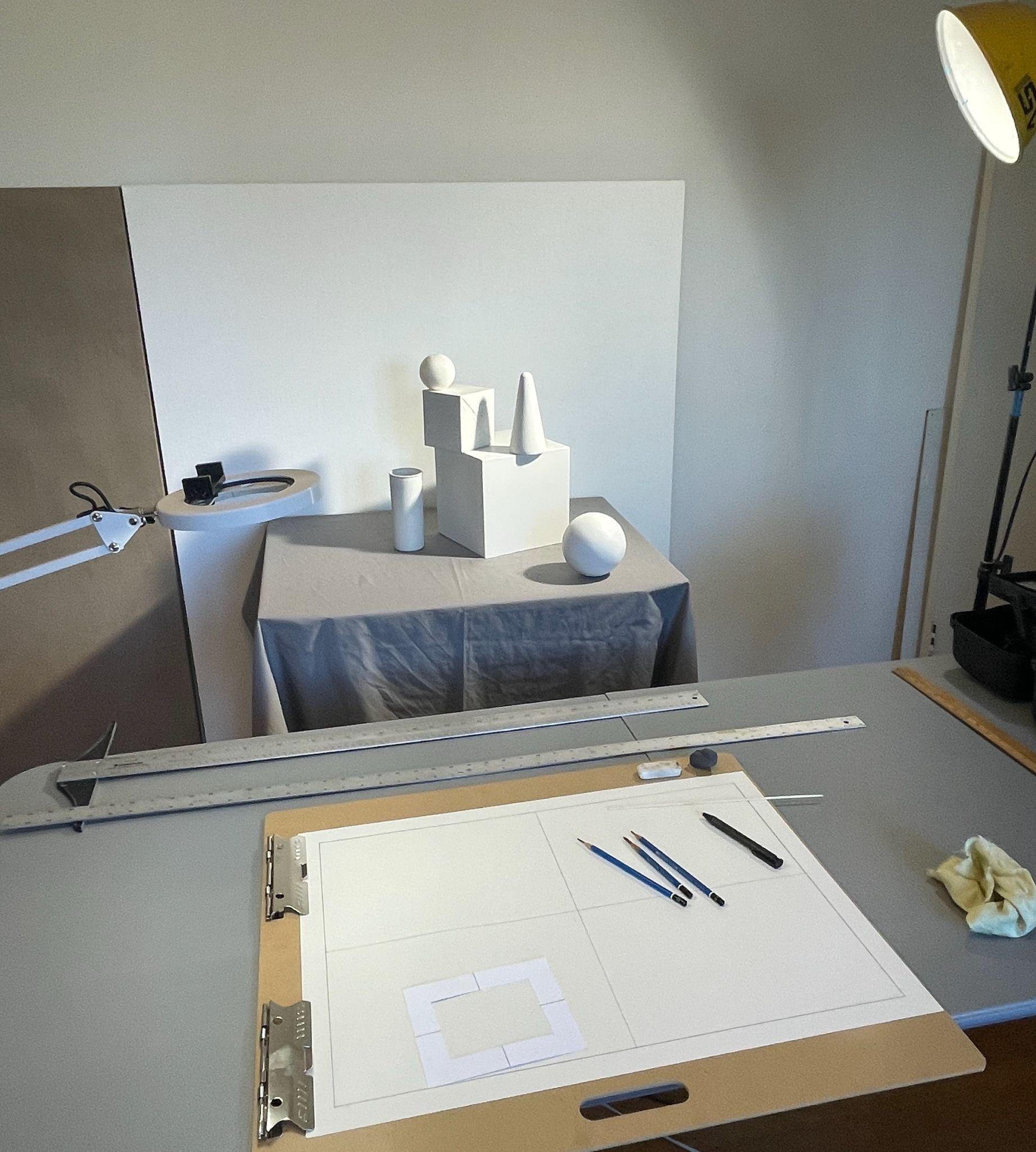 Photograph of an artist's workspace set up for still life drawing, featuring geometric shapes like a sphere, cone, cylinder, and cube arranged on a draped table. Workspace includes drawing tools such as pencils, pens, rulers, and a clipboard with divided paper, illuminated by two adjustable lamps.
