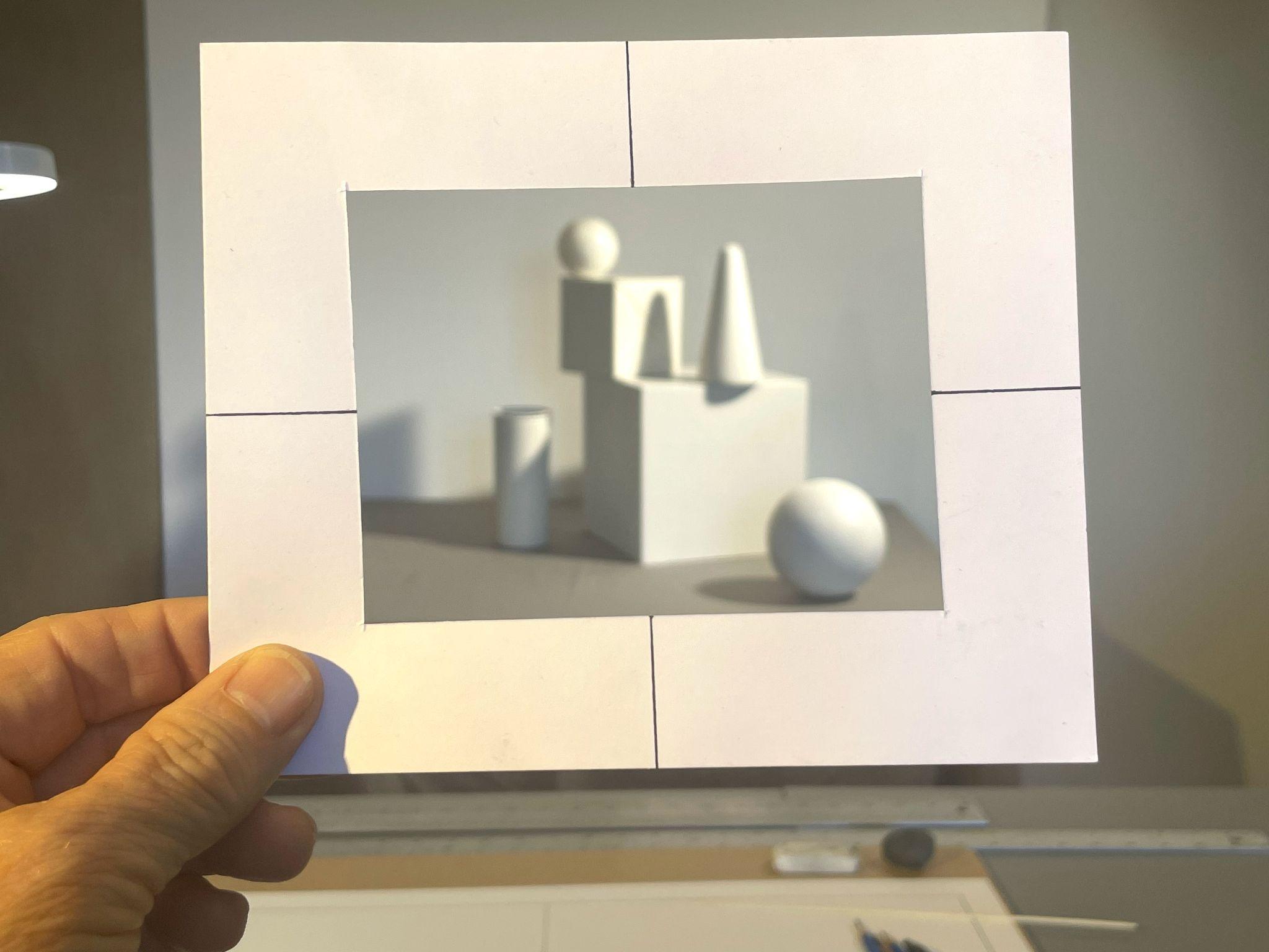 Photograph of a hand holding a white paper viewfinder frame with a rectangular cutout, focusing on a still life arrangement of white geometric shapes including a sphere, cone, cylinder, and cube. The composition highlights depth and shadow contrasts with a neutral background and visible drawing tools on a table below.