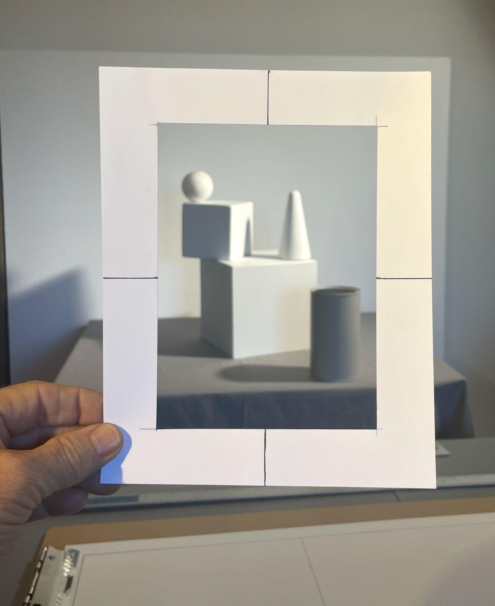 Photograph showing a hand holding a white paper viewfinder frame with black alignment marks, focusing on a still life arrangement of white geometric shapes including a sphere, cube, cone, and cylinder on a table. The setup highlights spatial relationships and perspective for drawing or painting practice, with soft lighting and neutral background.