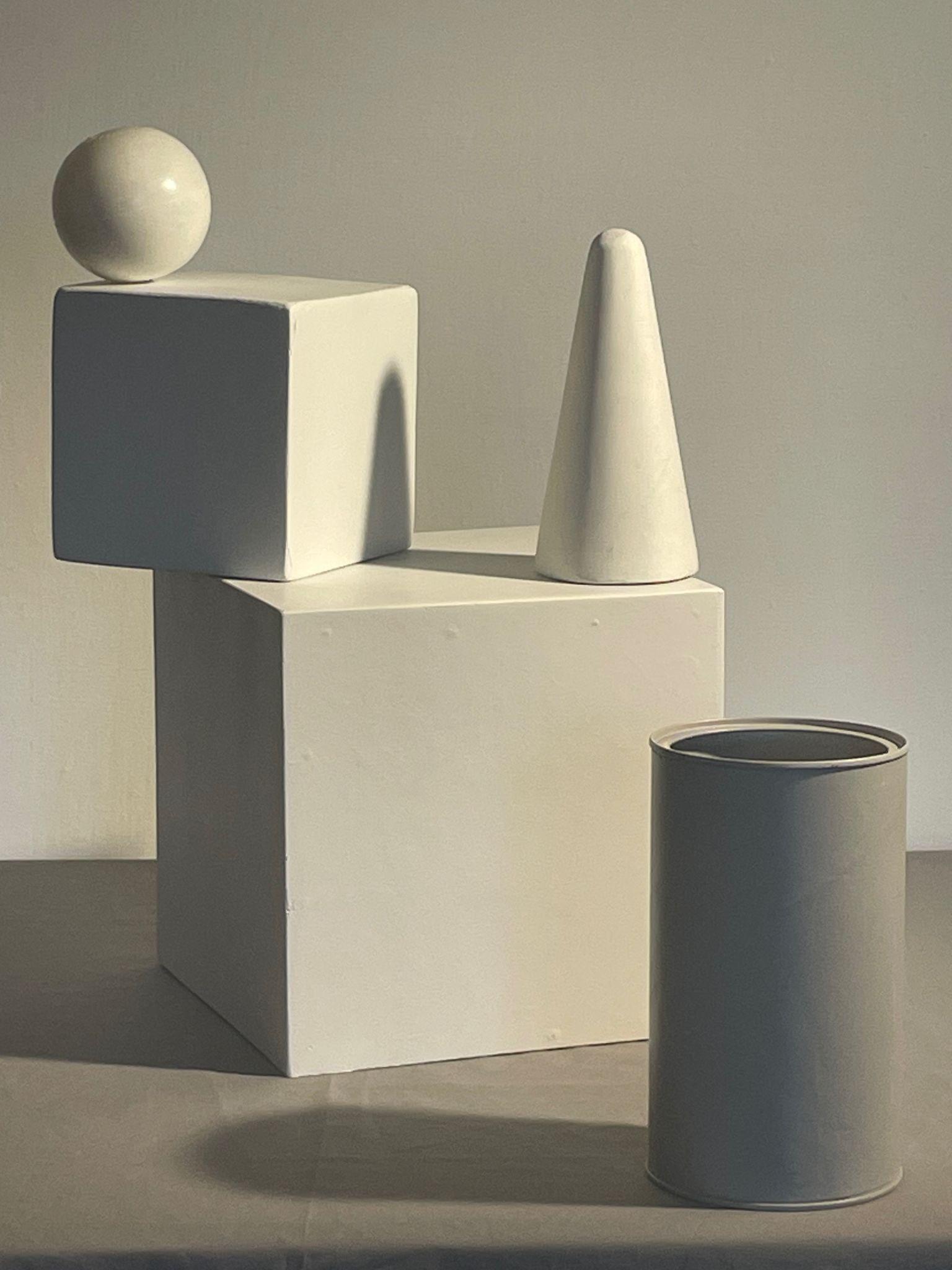 Photograph of a still life arrangement featuring four white geometric shapes: a sphere balanced on a cube, a cone, a larger cube, and a cylindrical container. Soft lighting casts shadows, emphasizing smooth surfaces and clean lines, creating a minimalist and balanced composition.