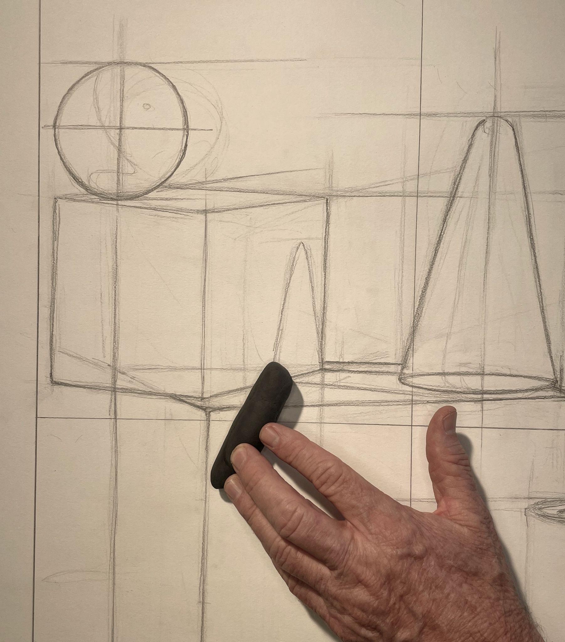 Photograph of a hand holding a black eraser over a pencil sketch of geometric shapes, including a sphere, cube, and cone, on a drawing surface. The sketch features construction lines and basic outlines, illustrating an early stage of a technical or artistic drawing process focused on form and perspective.