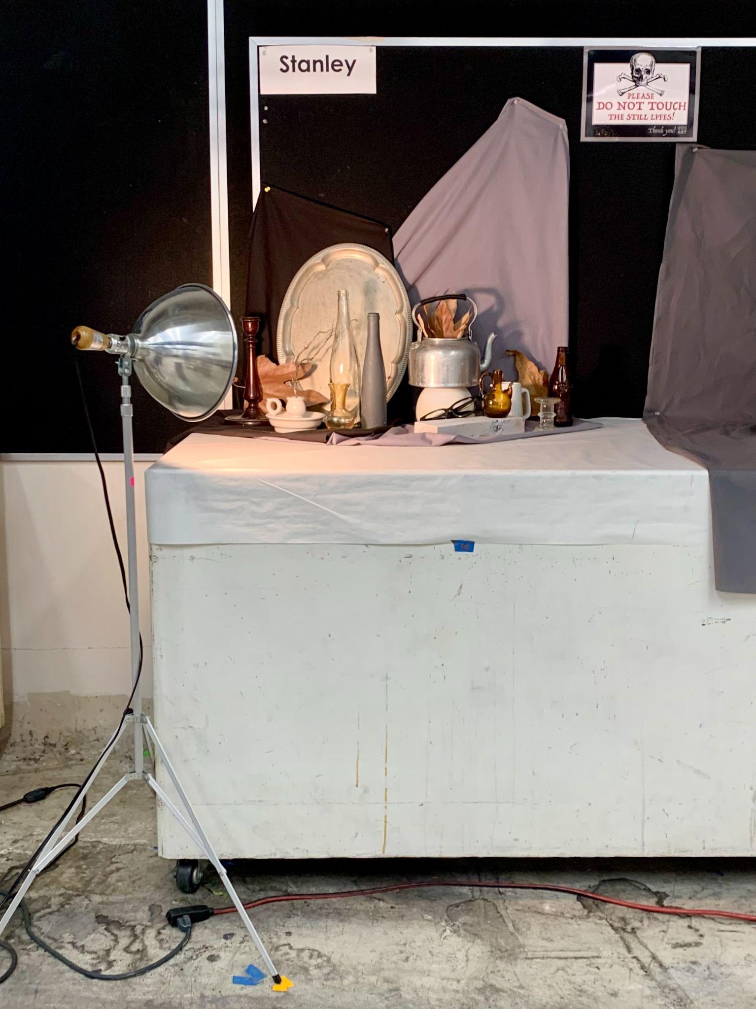 Photograph of a still life setup on a white table labeled "Stanley," featuring various objects such as a metal tray, glassware, a kettle, and bottles arranged against a black and gray backdrop. A large studio light on a tripod stands to the left, and a sign above warns "Do Not Touch," indicating a controlled work station.