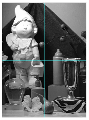Black and white photograph of a still life arrangement featuring a garden gnome figurine, a glass vase, a spray can, and a tall trophy cup on a patterned base. The composition includes decorative elements like a flower and spherical ornaments, creating a balanced display of varied textures and shapes.